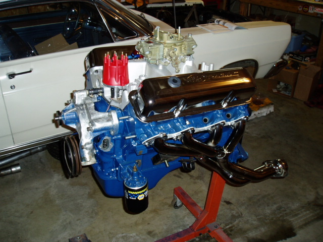 Rebuilt blue Ford V8 with chrome valve covers, carb, and headers on an engine stand in a garage