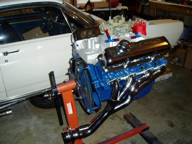 Blue Ford V8 with chrome valve covers, carburetor, and long-tube headers on a stand by a white car