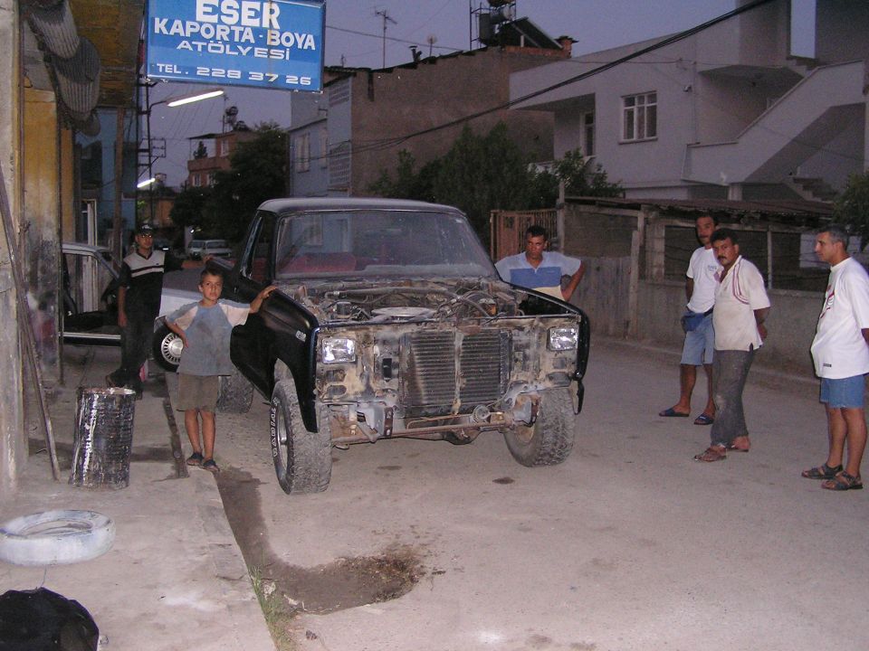Dark blue Ford truck with front grille and bumper removed at a Turkish body shop, surrounded by onlookers