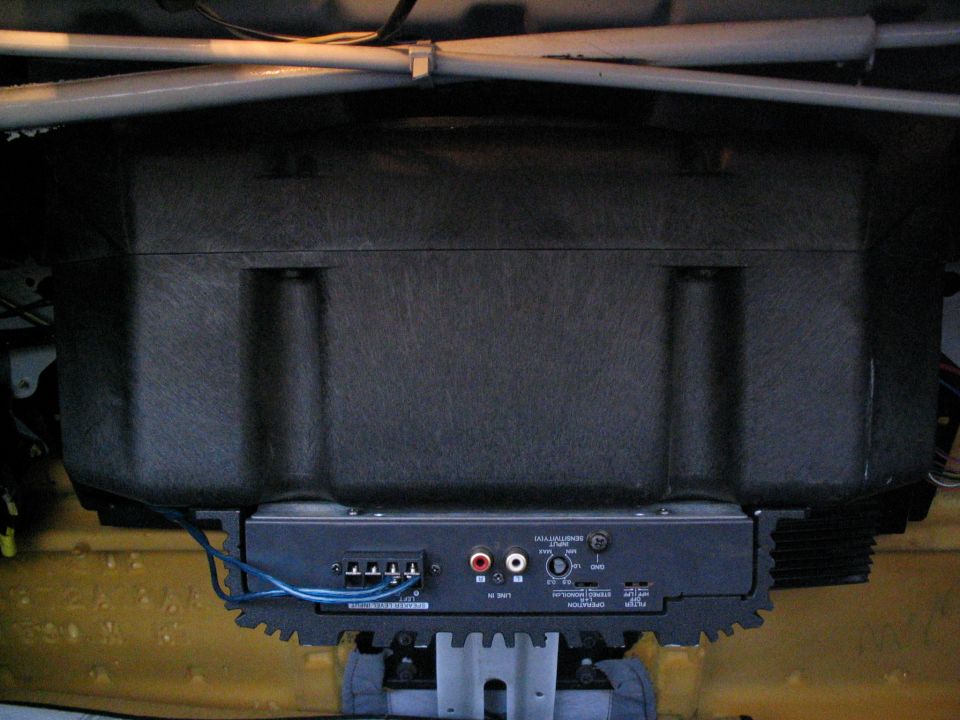 Car amplifier mounted under a carpeted subwoofer box in a trunk with blue wiring