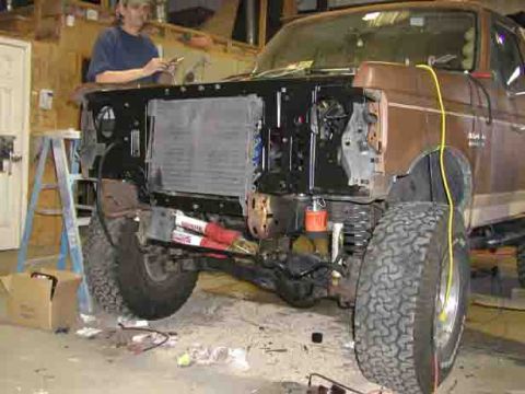 1988 Ford Bronco - Brown two-tone Ford 4x4 up in a shop with the grille, bumper and front clip stripped off, exposing the radiator and A/C condenser stack in…