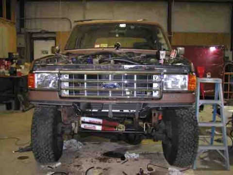 1988 Ford Bronco - Head-on view of a lifted brown/tan Ford 4x4 sitting in a shop with its front bumper and lower valance removed, exposing the frame horns, du…
