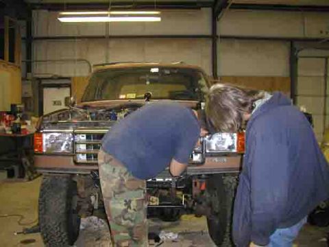 1988 Ford Bronco - Two individuals attend to the front of a brown Ford Bronco in a garage setting.