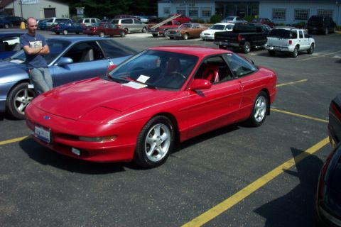 1993 Ford Probe - A red 1993 Ford Probe is parked in a lot, with other vehicles and a building in the background.
