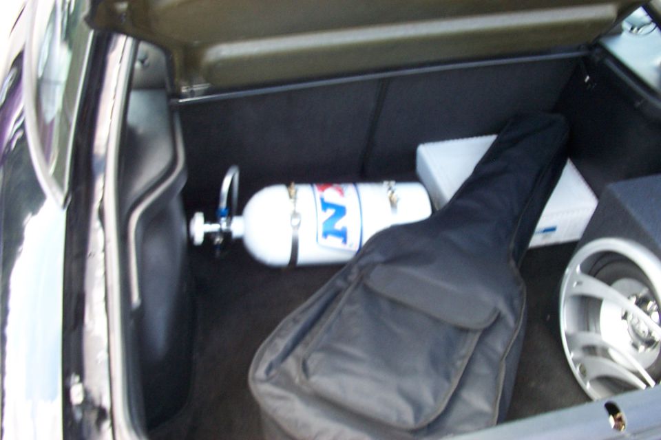 Car trunk holding a white nitrous bottle with blue lettering, a black guitar case, and a white box