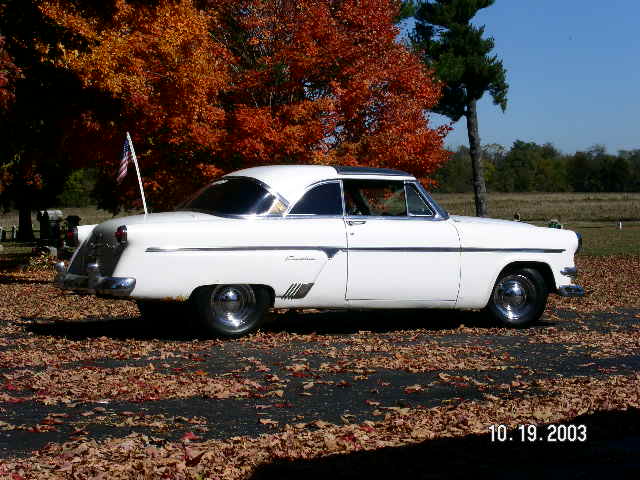 White 1950s Ford Crestline two-door hardtop parked among fall leaves, side view