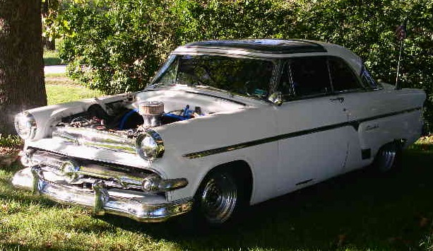 White 1950s Ford Crestline hardtop on grass with hood off showing blue engine and chrome air cleaner
