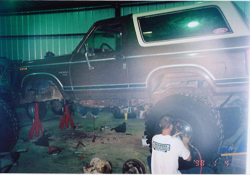 Brown Ford Bronco on jack stands mid lift install, man working at rear wheel in shop