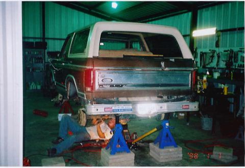 1984 Ford Bronco - A mechanic works on the rear of a brown Ford Bronco with a white top, supported by jacks and jack stands in a workshop.