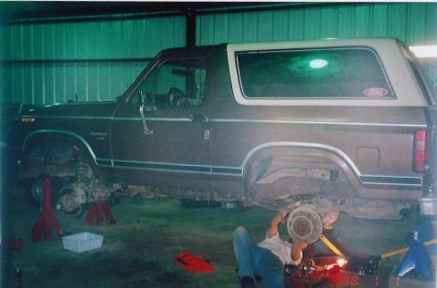 1984 Ford Bronco - A person works on the undercarriage of a 1984 Ford Bronco in a garage.