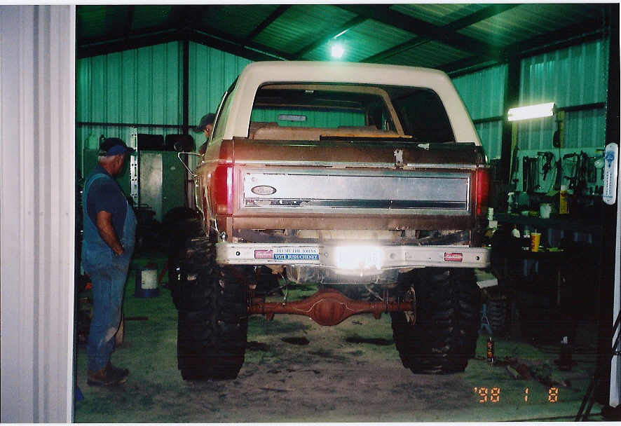 Rear of lifted brown Ford Bronco on mud tires inside a shop, man in overalls at left