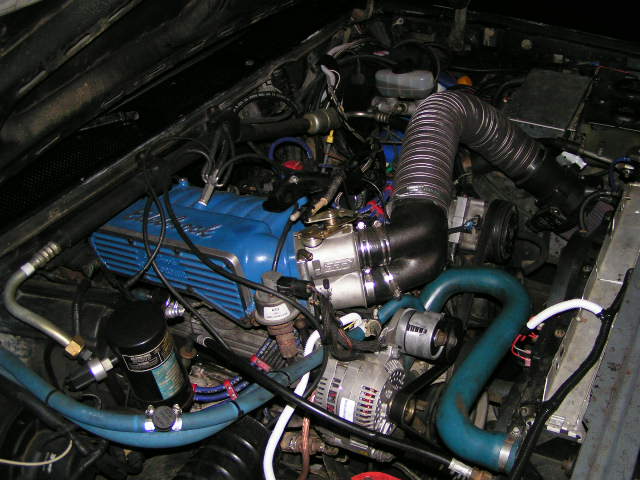 Ford engine with blue valve cover, ribbed intake duct, alternator and braided lines in bay