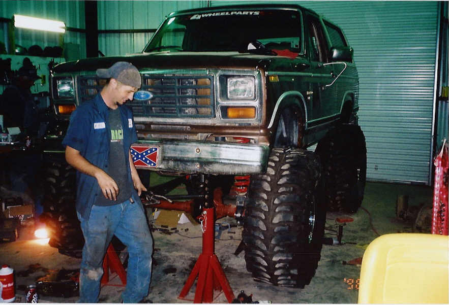 Green lifted Ford Bronco on jack stands with front suspension exposed in a shop