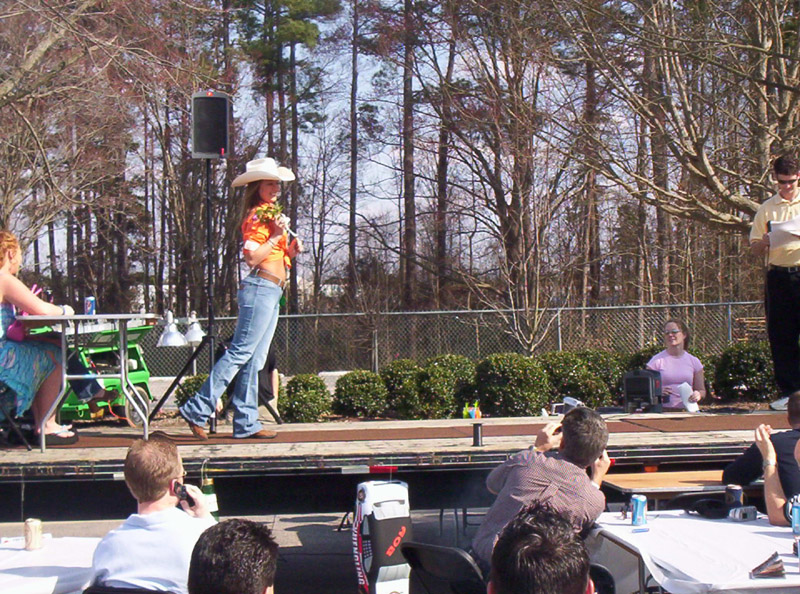 Outdoor event with a woman in a cowboy hat on a stage, audience seated at tables