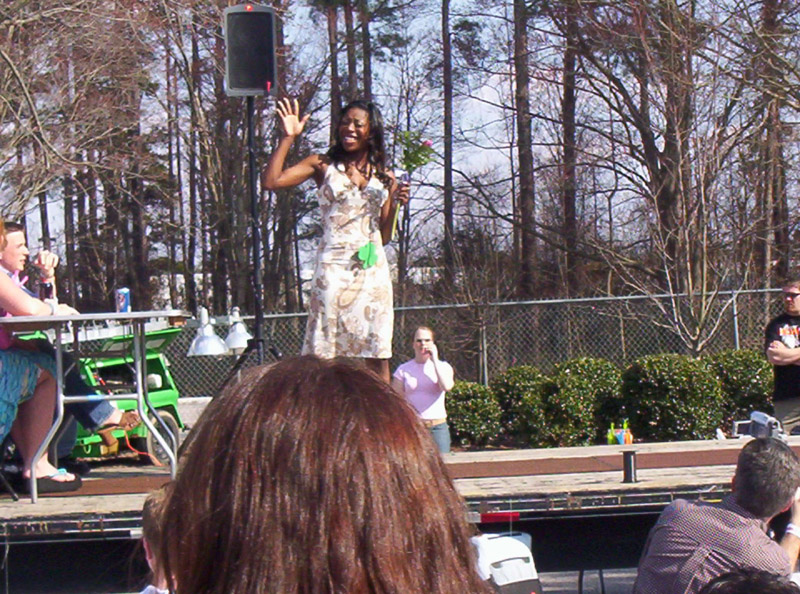 Woman in a patterned dress waves on a stage with trees and a fence in the background