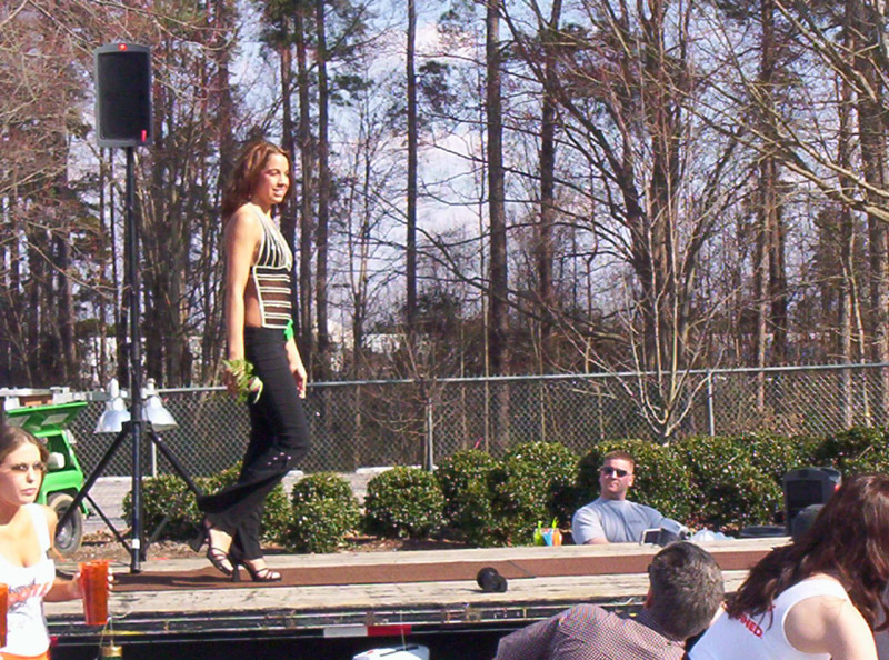 Woman in a striped top and black pants walks on a stage with trees and a fence in the background
