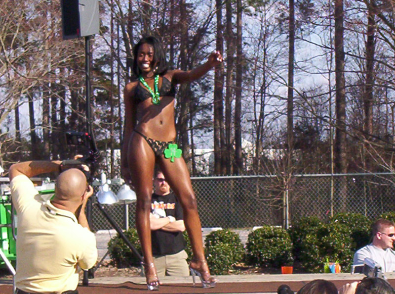 Woman in black bikini and heels posing on outdoor stage with photographer crouched at left