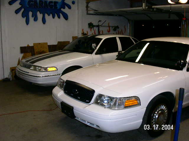 Two white Ford Crown Victoria police cars parked in a garage. One has black racing stripes on the hood