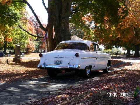 1954 Ford Crestline - A white 1954 Ford Crestline sedan is parked on a road covered in autumn leaves, with a cemetery in the background.