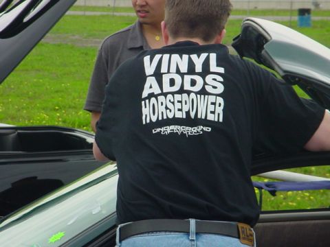 1978 Ford Mustang II - Two men stand beside a light-colored car with its hood raised at an outdoor grassy venue; the man facing away wears a black T-shirt reading…