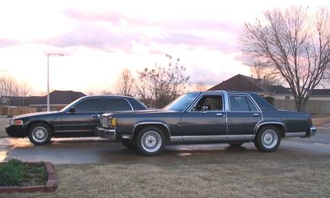1985 Ford Crown Victoria LTD - Two dark sedans, one identified as a 1985 Ford Crown Victoria LTD, are parked next to each other on a paved surface.