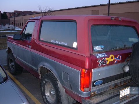 1995 Ford Bronco - Driver-side rear three-quarter view of a lifted two-tone red-over-silver full-size Ford Bronco in a snow-dusted parking lot, with a 'United…