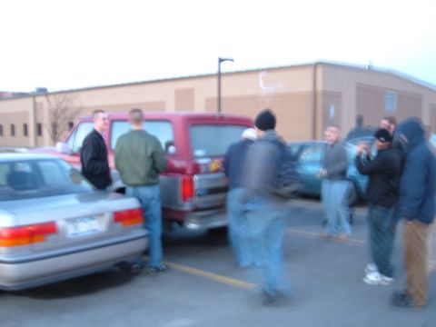 1995 Ford Bronco - Blurry dusk parking-lot gathering with a maroon full-size Ford Bronco parked nose-in beside a silver sedan, seen from the rear quarter with…
