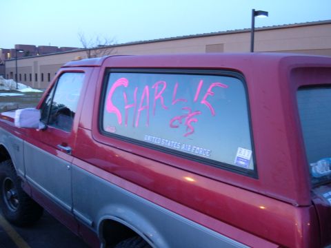 1995 Ford Bronco - Close side view of the rear quarter of a red-over-silver two-tone 1990s Ford Bronco parked at dusk in a lot beside a tan commercial buildin…