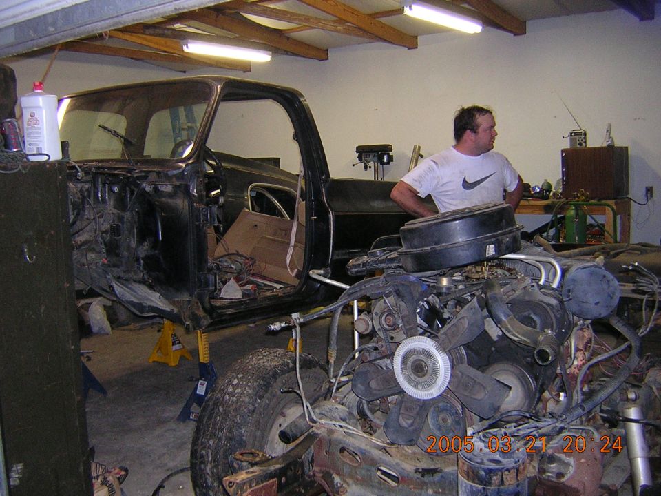 Stripped dark truck body on jack stands with V8 engine in foreground and man in garage