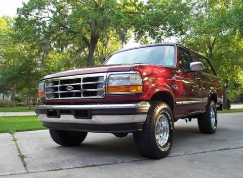 1996 Ford Bronco - Low three-quarter front view of a dark red two-door Ford Bronco parked on a driveway under Spanish-moss-draped oaks, with chrome grille, bu…