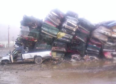 1971 Ford Mustang - A large pile of wrecked vehicles, with a single white pickup truck in the foreground, sits in a muddy junkyard.