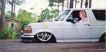 1990 Ford Bronco - white full-size Ford Bronco dropped flat on the pavement with billet wheels and a woman seated at the wheel near a shed.