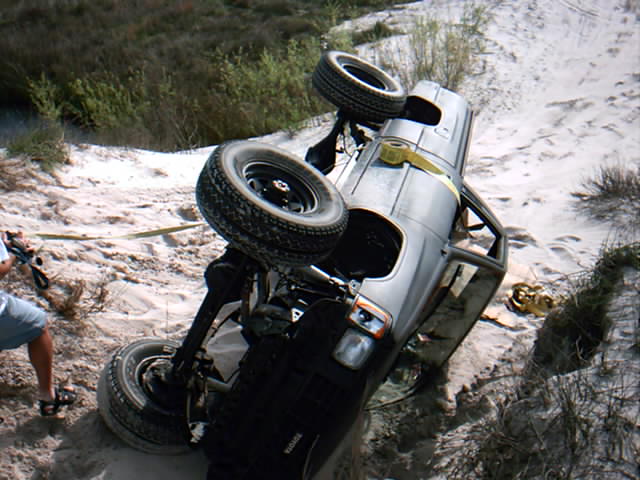 Silver truck rolled onto its side in sand dunes with yellow recovery strap and wheels in the air
