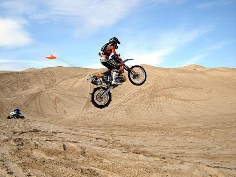 1990 Ford Bronco - helmeted dirt bike rider launches off a sand dune jump while a quad waits below under a blue sky.