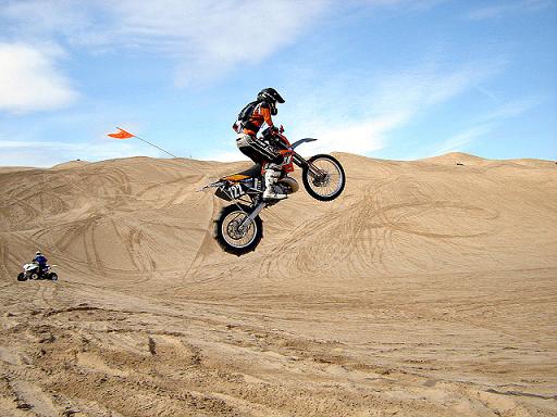 Dirt bike rider catching air over sand dunes with an ATV in the background