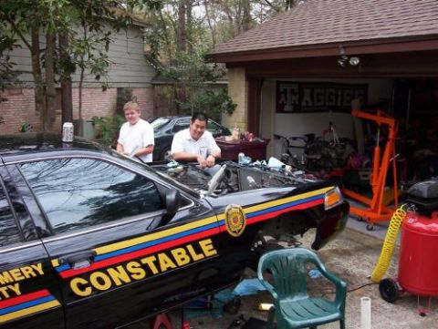 2000 Ford Crown Victoria - Two men work on the engine of a black police car with 'CONSTABLE' text on its side, parked in a driveway next to a garage.