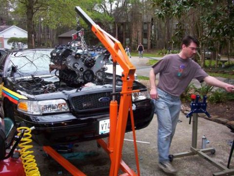 2000 Ford Crown Victoria - An engine is being lifted out of a black car with an engine hoist. A man is overseeing the process.