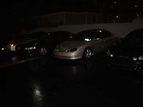 1993 Ford Taurus - A silver sedan is parked in a lot at night, with other vehicles nearby.