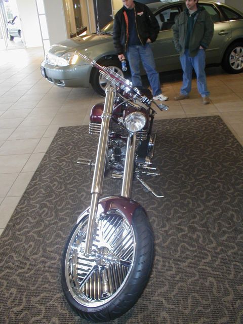 1979 Ford Bronco - A custom chopper motorcycle is displayed indoors with two men and a car visible behind it.