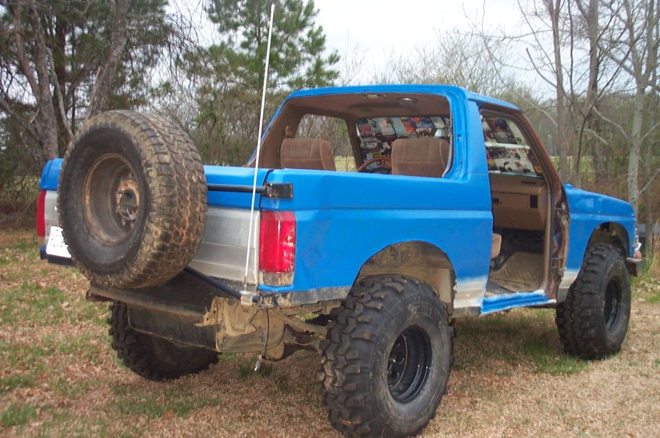 Blue Ford Bronco with a spare tire on the back, lifted with large tires, and the driver's door removed