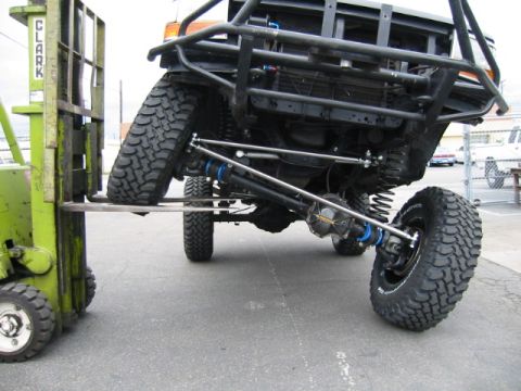 1992 Ford Bronco - Front suspension flex test with one wheel lifted by the forks of a green Clark forklift, showing the coil-sprung solid front axle, radius a…