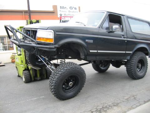 1992 Ford Bronco - Black lifted Ford Bronco flexing its front suspension while lifted by a green forklift in a parking lot, front driver wheel drooped far bel…