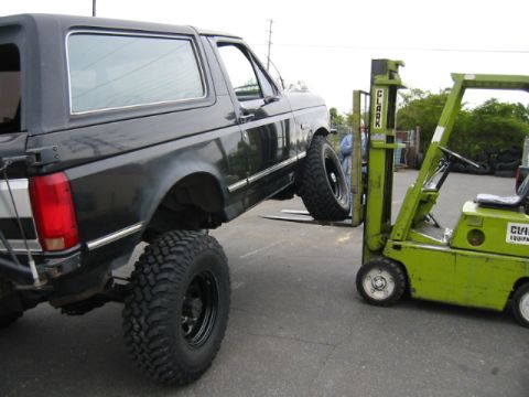 1992 Ford Bronco - A lifted black Ford Bronco flexes its suspension in a parking lot with its driver-side front wheel ramped up on the forks of a green Clark…