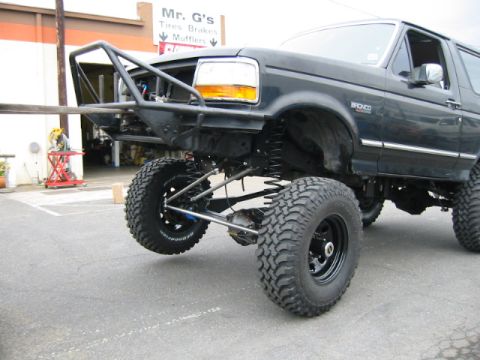 1992 Ford Bronco - Front three-quarter view of a lifted dark Ford Bronco articulating at max droop, one front wheel hanging low to show the coil-sprung solid…