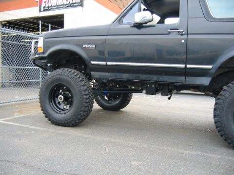 1992 Ford Bronco - Low side view of a lifted dark-colored Ford Bronco showing ride height, with black steel wheels, tall mud-terrain tires, exposed coil-sprun…