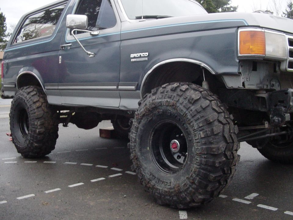 Lifted blue Ford Bronco on oversized mud tires, BRONCO fender badge, parked on wet asphalt