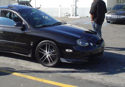 1993 Ford Taurus - A black sedan with aftermarket wheels is parked in a lot, with another car behind it.