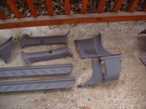 1993 Ford Probe - Gray plastic interior trim panels laid out on a concrete patio beside a wooden deck rail: a pair of A-pillar covers with belt-guide holes,…