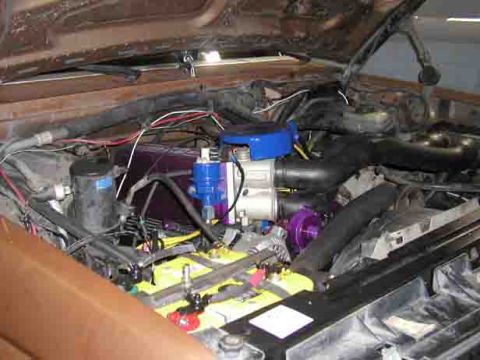 1988 Ford Bronco - Open hood view down into a crowded engine bay with a purple-anodized intake and throttle body, blue-painted air-intake elbow and fuel-rail…