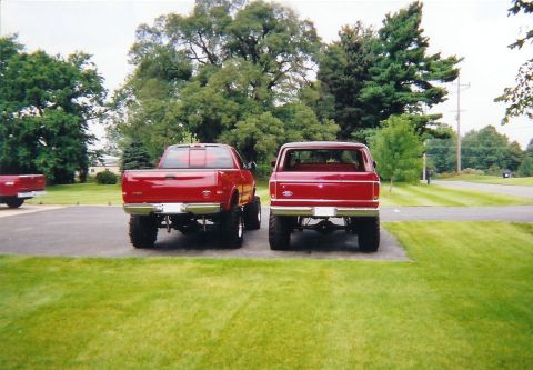 1978 Ford Bronco - Two red pickup trucks are parked in a grassy area, viewed from the rear.